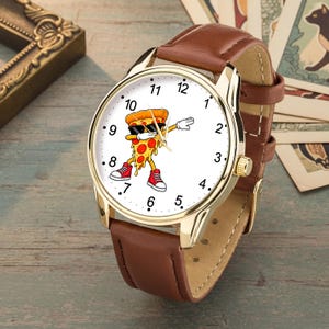 May include: A wristwatch with a gold-tone metal case and a brown leather strap. The white watch face features a graphic of a cartoon pizza slice wearing black sunglasses and red sneakers, performing a dabbing pose, surrounded by black numerals.