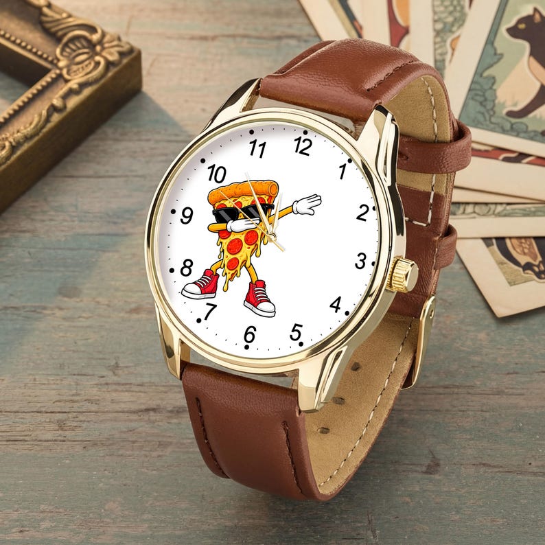 May include: A wristwatch with a gold-tone metal case and a brown leather strap. The white watch face features a graphic of a cartoon pizza slice wearing black sunglasses and red sneakers, performing a dabbing pose, surrounded by black numerals.