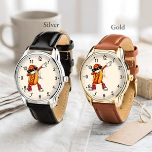 May include: Two analog wristwatches with cream-colored faces featuring a cartoon hot dog character dabbing. The left watch has a silver-tone metal case and a black leather strap. The right watch has a gold-tone metal case and a brown leather strap.