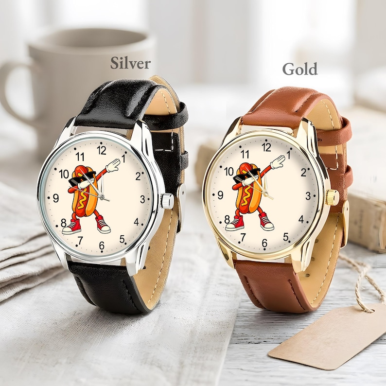 May include: Two analog wristwatches with cream-colored faces featuring a cartoon hot dog character dabbing. The left watch has a silver-tone metal case and a black leather strap. The right watch has a gold-tone metal case and a brown leather strap.
