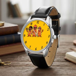 May include: A wristwatch with a black leather strap and a silver-tone metal case. The watch face is bright yellow and features a graphic illustration of three cartoon hot dogs wearing sunglasses and hats, with black numerals marking the hours.