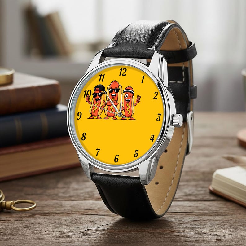 May include: A wristwatch with a black leather strap and a silver-tone metal case. The watch face is bright yellow and features a graphic illustration of three cartoon hot dogs wearing sunglasses and hats, with black numerals marking the hours.