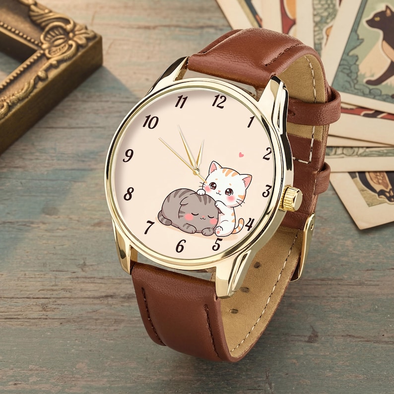 May include: A wristwatch with a gold-tone metal case and a brown leather strap. The watch face is cream-colored with black numerals and features a graphic illustration of two small, cartoon-style cats cuddling together in the center.