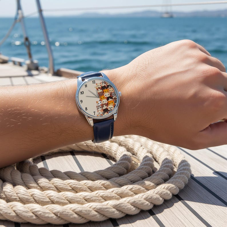 May include: A wristwatch with a silver case and a navy blue leather band. The watch face features a collection of cartoon cat faces. The watch is being worn on a person's wrist.