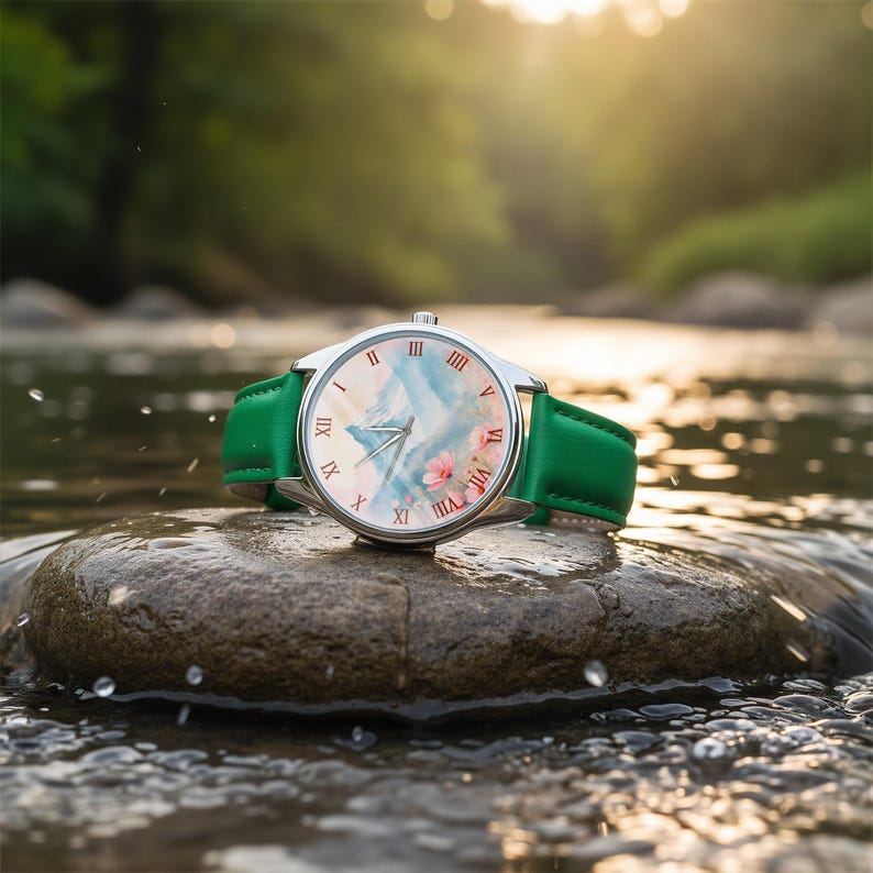 K&ouml;nnte beinhalten: Eine silberne Armbanduhr mit gr&uuml;nem Lederarmband liegt auf einem Felsen in einem Bach. Das Zifferblatt der Uhr zeigt eine Berglandschaft mit rosa Blumen und r&ouml;mischen Ziffern. Ein stilvolles Accessoire.