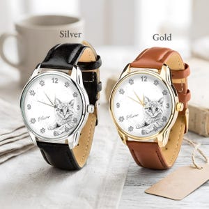 May include: Two wristwatches with cat-themed faces. One watch has a silver case and a black leather band, the other has a gold case and a brown leather band. The watch faces feature a cat illustration, paw prints, and the name "Oliver".