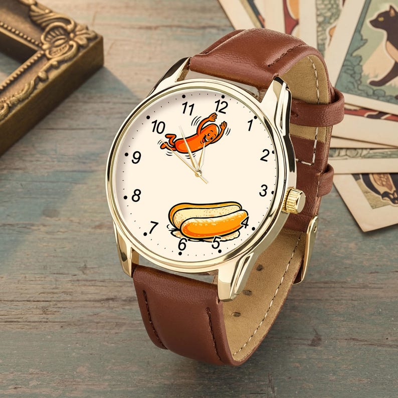 May include: A gold-tone wristwatch with a brown leather strap and a cream-colored dial. The watch face features black numerals from 1 to 12, a cartoon hot dog character near the 12 o'clock position, and a hot dog illustration near the 5 o'clock mark.