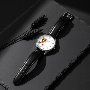 May include: A wristwatch with a black leather strap and a silver-tone case. The white watch face features black numerals from 1 to 12 and a central graphic of a cartoon pizza slice wearing sunglasses and red sneakers, positioned in a dynamic pose.
