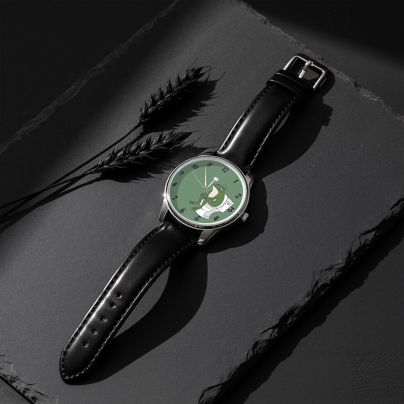 May include: A wristwatch with a black leather band and a silver-colored case. The watch face is green with a cartoon frog design. The watch is lying on a dark surface.