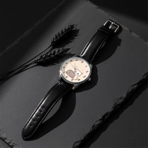 May include: A wristwatch with a black leather strap and a silver-tone circular case. The watch face is cream-colored and features a graphic illustration of two small cats, one grey and one orange, with black numerals and thin silver hands.