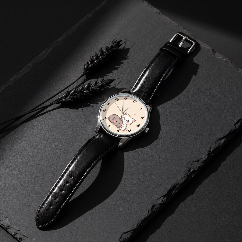 May include: A wristwatch with a black leather strap and a silver-tone circular case. The watch face is cream-colored and features a graphic illustration of two small cats, one grey and one orange, with black numerals and thin silver hands.