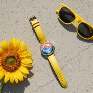 May include: A yellow wristwatch with a mountain landscape dial and a matching yellow leather strap. The watch is next to a pair of yellow sunglasses and a sunflower. The watch face features a sunrise over a mountain range.
