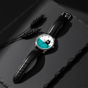 Pu&ograve; includere: Un orologio da polso con cinturino in pelle nera e cassa color argento. Il quadrante presenta un design stravagante di un gatto nero in una scena acquatica turchese con piccoli pesci arancioni. Le lancette indicano l'ora.