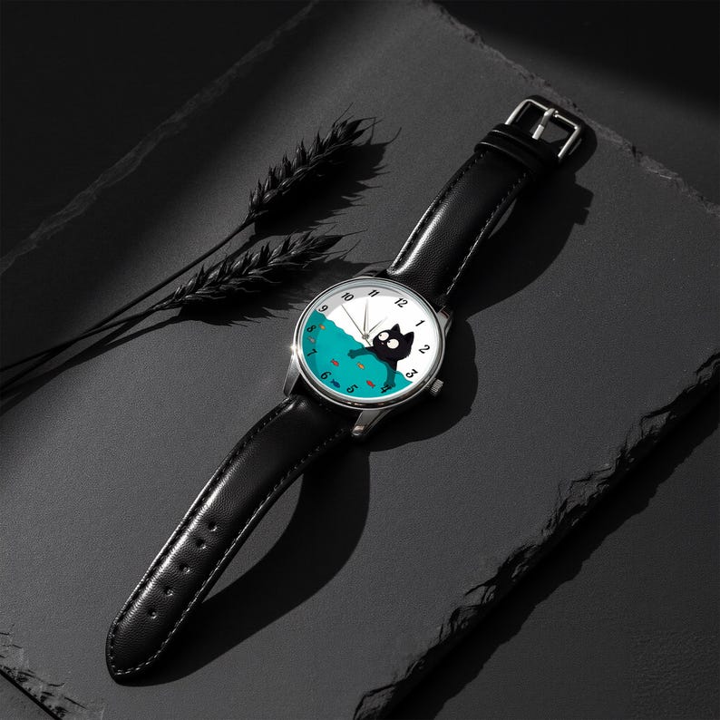 Pu&ograve; includere: Un orologio da polso con cinturino in pelle nera e cassa color argento. Il quadrante presenta un design stravagante di un gatto nero in una scena acquatica turchese con piccoli pesci arancioni. Le lancette indicano l'ora.