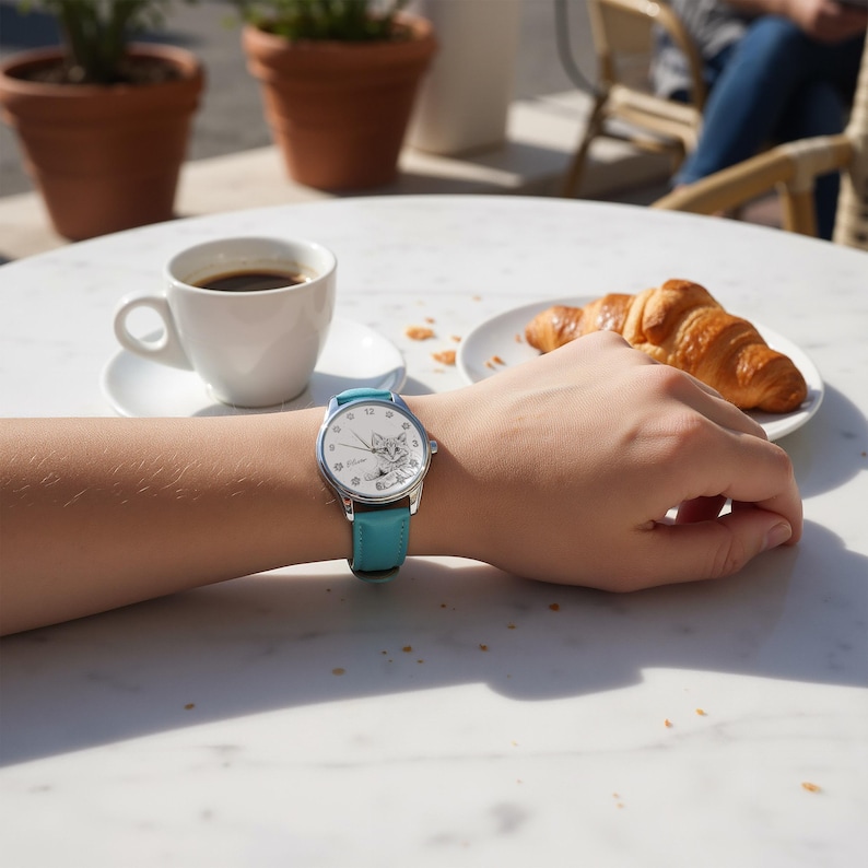 May include: A silver wristwatch with a light blue leather band. The watch face features a cartoon cat illustration and white numerals. The watch is on a person's wrist, with a cup of coffee and a croissant in the background.