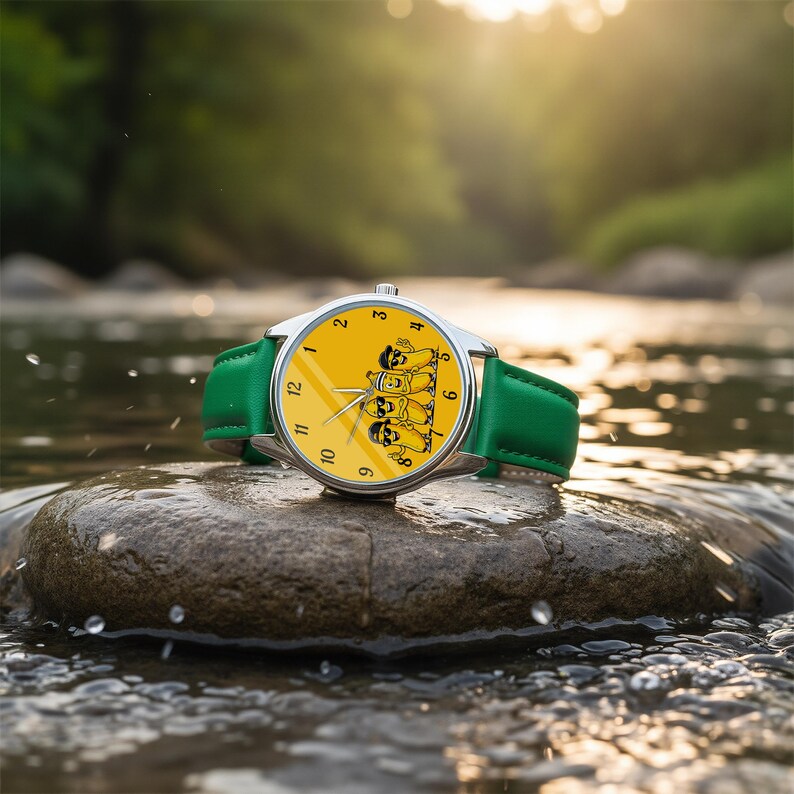 Puede incluir: Un reloj de pulsera con una esfera de color amarillo brillante que presenta un gr&aacute;fico de cuatro pl&aacute;tanos de dibujos animados con gafas de sol. El reloj tiene una caja de metal plateado y una correa de cuero verde liso con n&uacute;meros negros.