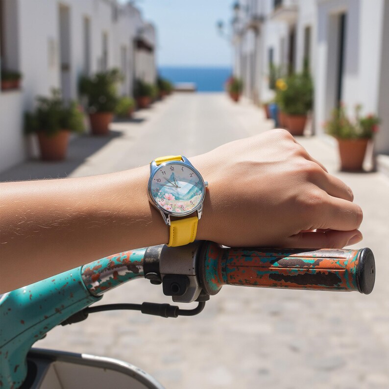 May include: A wristwatch with a bright yellow leather strap and a silver-tone metal case. The watch face features a light blue and white graphic illustration of a wave with small pink floral accents and black numerical hour markers.