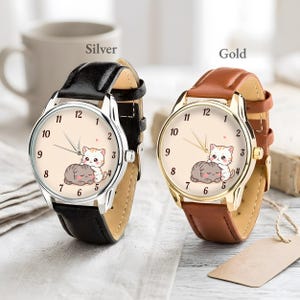 May include: Two analog wristwatches with round faces featuring a cartoon illustration of two cats. The left watch has a silver-tone case and a black leather strap. The right watch has a gold-tone case and a brown leather strap. Both dials show numbers 1 through 12.