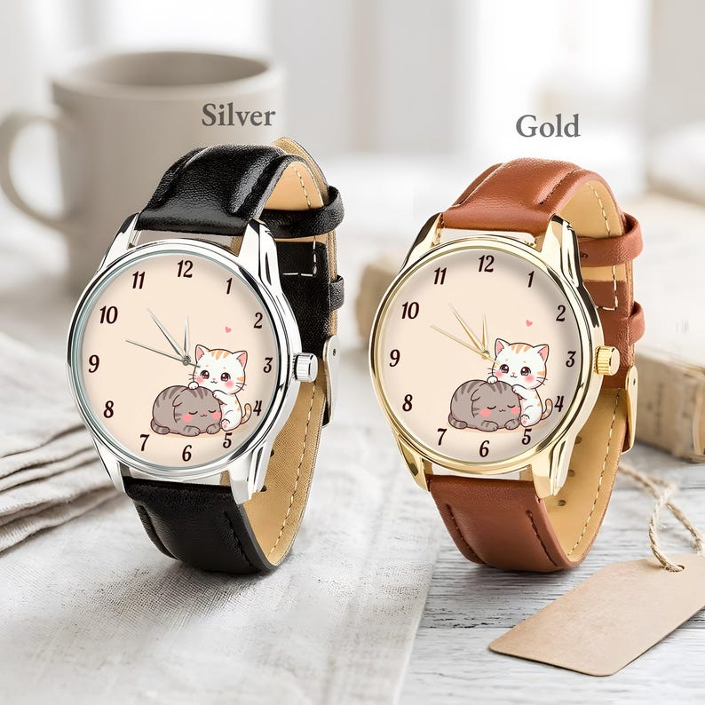 May include: Two analog wristwatches with round faces featuring a cartoon illustration of two cats. The left watch has a silver-tone case and a black leather strap. The right watch has a gold-tone case and a brown leather strap. Both dials show numbers 1 through 12.