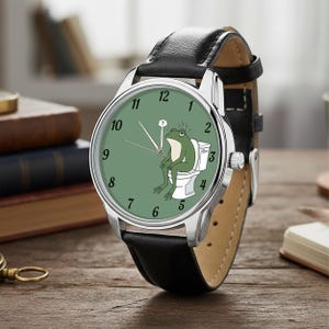 May include: A silver wristwatch with a black leather band. The watch face is green with white numerals and features a cartoon frog sitting on a toilet. The watch has a silver bezel and a brown leather backing.