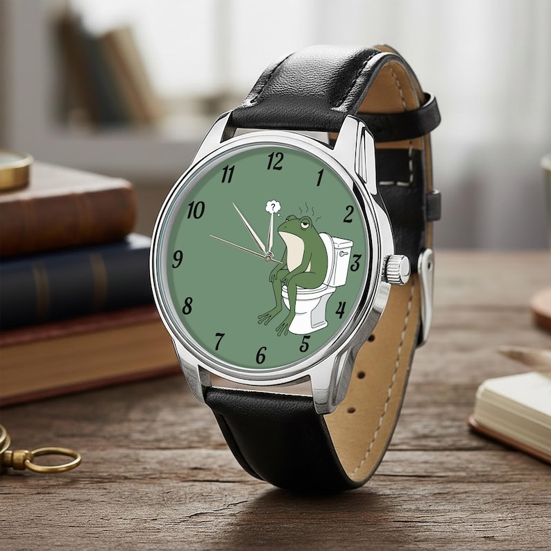 May include: A silver wristwatch with a black leather band. The watch face is green with white numerals and features a cartoon frog sitting on a toilet. The watch has a silver bezel and a brown leather backing.