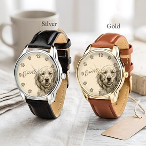 Pu&ograve; includere: Due orologi con cinturini in pelle. Uno ha una cassa argentata e un cinturino nero, l'altro una cassa dorata e un cinturino marrone. Entrambi gli orologi hanno un quadrante color crema con un ritratto di cane, impronte di zampe e il nome "Daisy".