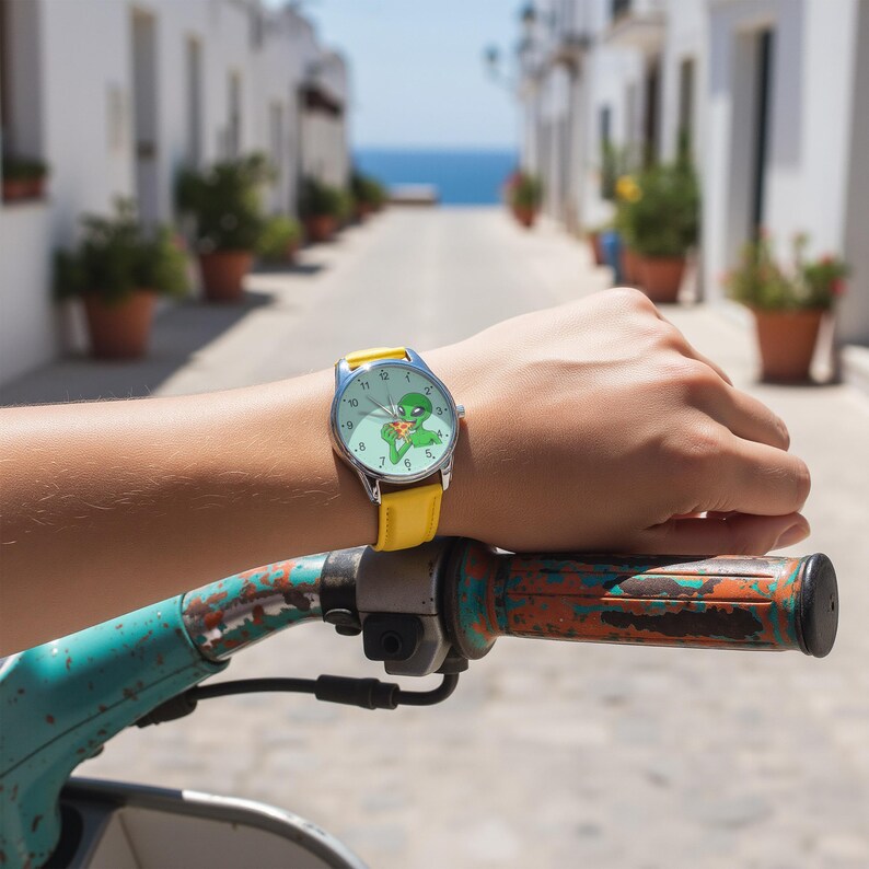 May include: A wristwatch with a bright yellow leather strap and a silver-tone metal case. The watch face features a light green background with a graphic illustration of a green alien eating a slice of pizza, complete with black hour markers.