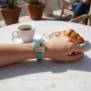 Puede incluir: Un reloj de pulsera con una correa de cuero azul claro y una caja plateada. La esfera del reloj presenta una colorida ilustraci&oacute;n de un gato y una persona. El reloj est&aacute; en una mu&ntilde;eca, con una taza de caf&eacute; y un cruas&aacute;n en el fondo.