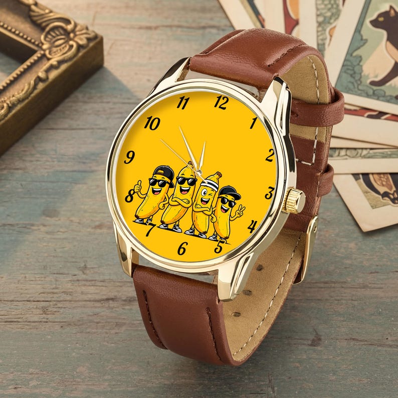 Puede incluir: Un reloj de pulsera con correa de cuero marr&oacute;n y caja de metal en tono dorado. La esfera es de color amarillo brillante y presenta una ilustraci&oacute;n gr&aacute;fica de cuatro pl&aacute;tanos de dibujos animados con gafas de sol y sombreros, con n&uacute;meros negros.