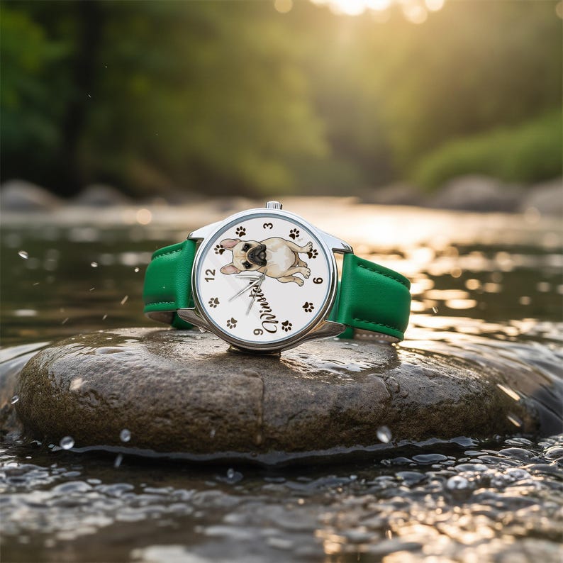 May include: A silver wristwatch with a white face featuring a pug illustration and the name "Winston". The watch has black paw prints and green leather band. The watch is resting on a rock in a stream.