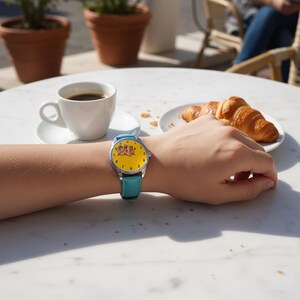 May include: A wristwatch with a bright yellow face featuring a small graphic of three cartoon characters, black numerals, and a silver-tone metal case. The watch is secured with a smooth, solid light blue leather strap with a silver buckle.