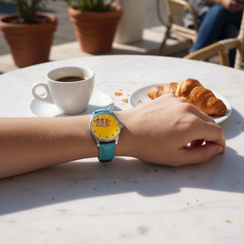 May include: A wristwatch with a bright yellow face featuring a small graphic of three cartoon characters, black numerals, and a silver-tone metal case. The watch is secured with a smooth, solid light blue leather strap with a silver buckle.