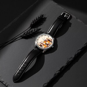 May include: A wristwatch with a black leather band and a silver-colored case. The watch face features a cream-colored background with a pattern of cartoon cats in various colors. The watch is displayed on a dark surface.