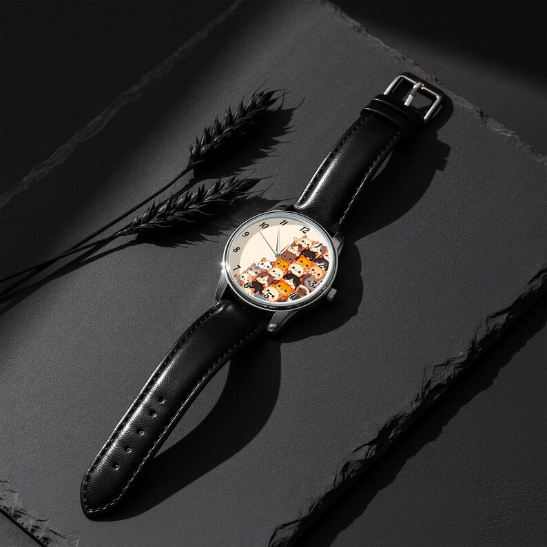 May include: A wristwatch with a black leather band and a silver-colored case. The watch face features a cream-colored background with a pattern of cartoon cats in various colors. The watch is displayed on a dark surface.