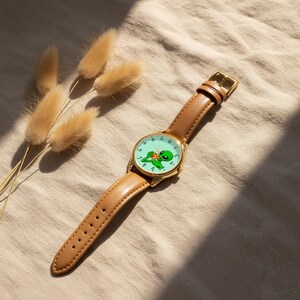 May include: A wristwatch with a tan leather strap and a gold-tone metal case. The watch face is light green and features a graphic illustration of a green alien eating a slice of pizza, with black numbers marking the hours around the perimeter.