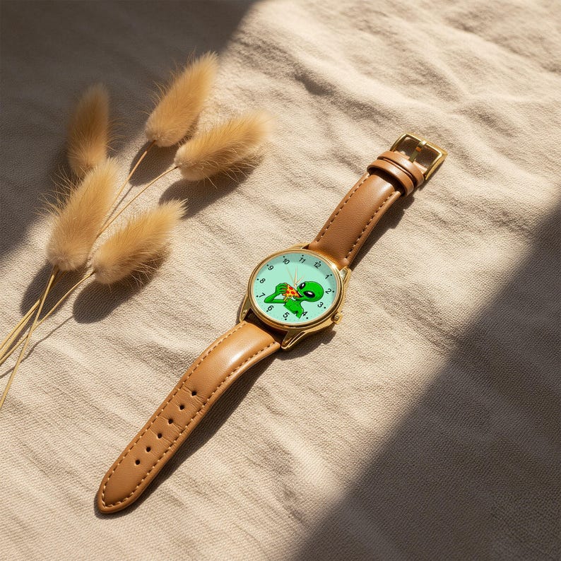 May include: A wristwatch with a tan leather strap and a gold-tone metal case. The watch face is light green and features a graphic illustration of a green alien eating a slice of pizza, with black numbers marking the hours around the perimeter.