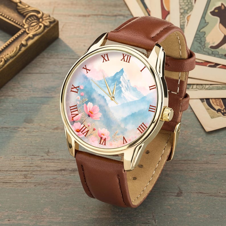 K&ouml;nnte beinhalten: Goldfarbene Armbanduhr mit braunem Lederarmband. Das Zifferblatt zeigt eine Berglandschaft mit rosa Bl&uuml;ten und r&ouml;mischen Ziffern. Die Uhr hat eine goldene L&uuml;nette und ein braunes Lederarmband.