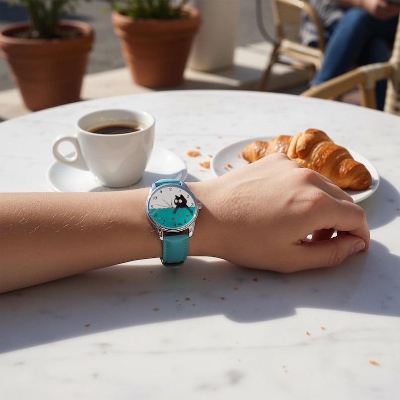 Pu&ograve; includere: Orologio da polso rotondo con cinturino in pelle azzurra. Il quadrante dell'orologio presenta un gatto nero su sfondo turchese, con numeri e lancette bianche. L'orologio &egrave; al polso, con una tazza di caff&egrave; e un croissant sullo sfondo.