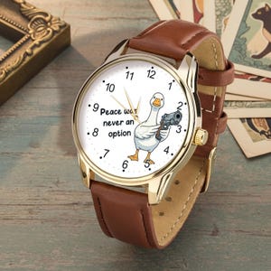 Peut inclure: Montre-bracelet dorée avec un bracelet en cuir marron. Le cadran de la montre présente un dessin d'oie tenant un pistolet, avec le texte "Peace was never an option". Le cadran est blanc avec des chiffres noirs.