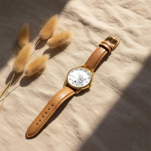 May include: A gold-tone wristwatch with a light brown leather band. The watch face features a white background with a black and white illustration of a cat. The watch is set against a neutral-toned background with dried flowers.