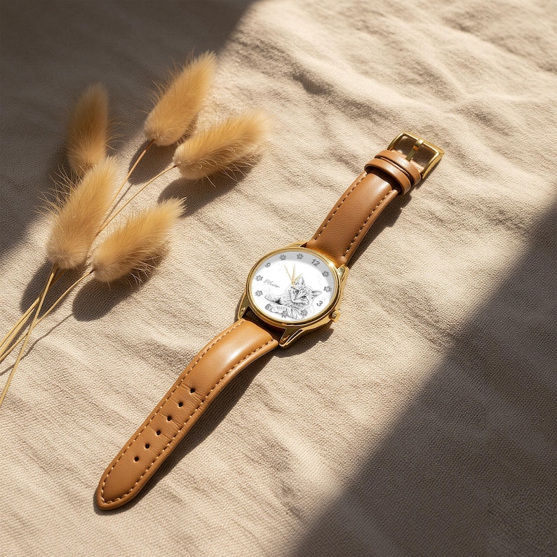 May include: A gold-tone wristwatch with a light brown leather band. The watch face features a white background with a black and white illustration of a cat. The watch is set against a neutral-toned background with dried flowers.