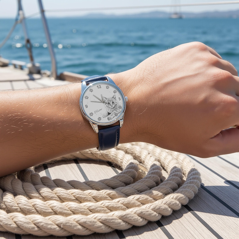 May include: A silver wristwatch with a white face featuring a cat illustration and the name "Elaine". The watch has a navy blue leather band. The watch is on a person's wrist.