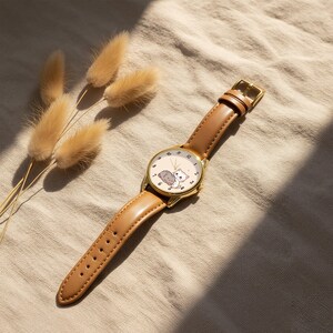 May include: A wristwatch with a gold-tone circular case and a tan leather strap. The watch face is white with black numerals and features a small illustration of a white cat and a grey cat sitting together in the center of the dial.