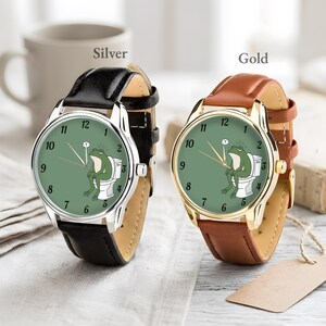 May include: Two wristwatches, one silver and one gold, each with a leather band. The watch faces are green with black numbers and hands. A cartoon frog is sitting on a toilet on the watch face. The word "Silver" and "Gold" are above the watches.