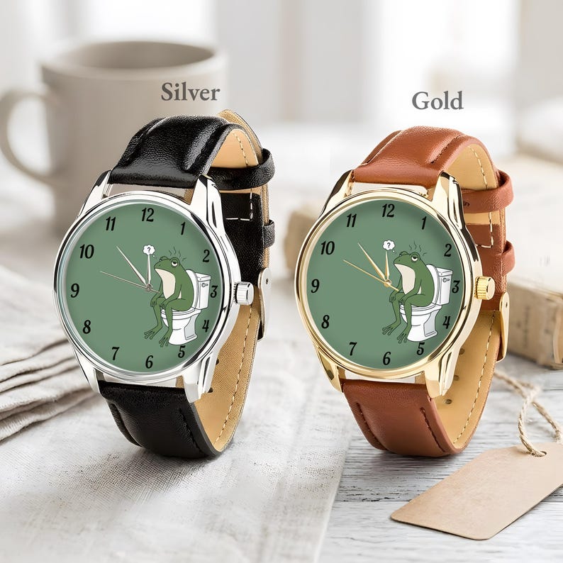 May include: Two wristwatches, one silver and one gold, each with a leather band. The watch faces are green with black numbers and hands. A cartoon frog is sitting on a toilet on the watch face. The word "Silver" and "Gold" are above the watches.