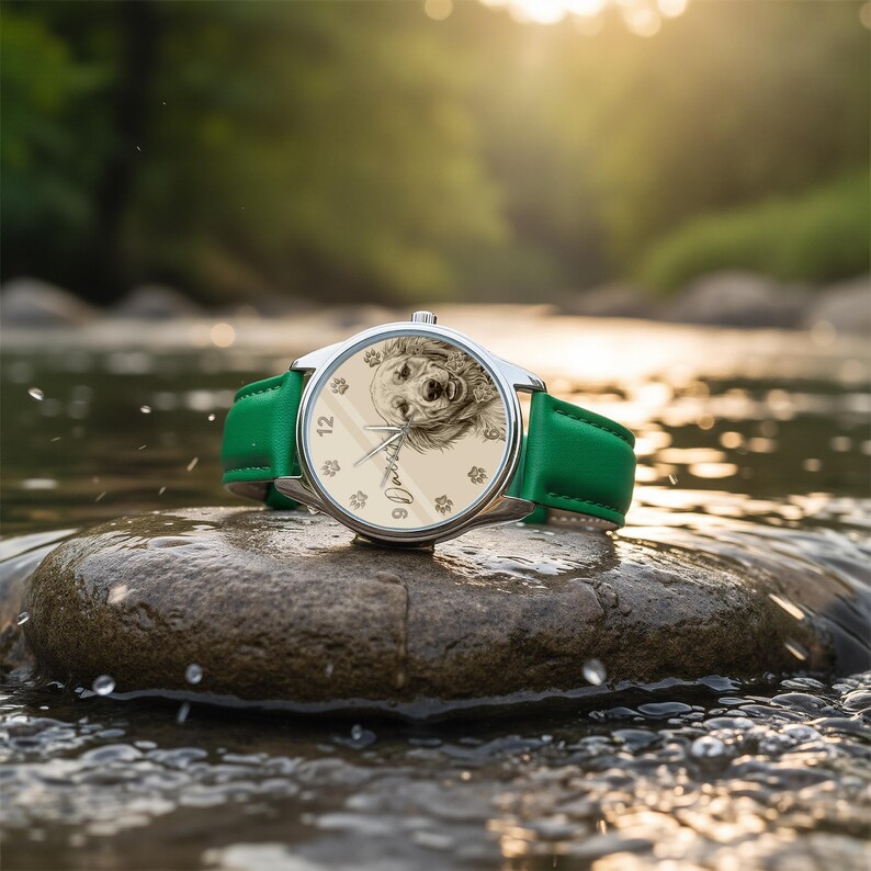 Pu&ograve; includere: Un orologio da polso argentato con cinturino in pelle verde &egrave; appoggiato su una roccia in un ruscello. Il quadrante dell'orologio presenta un'illustrazione dettagliata del volto di un cane, con il nome "Dakota" scritto sotto. Il quadrante &egrave; color crema con numeri neri.
