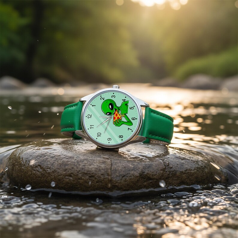 May include: A wristwatch with a silver-toned metal case and a green leather strap. The watch face is light green and features a graphic illustration of a green alien eating a slice of pepperoni pizza, with black numerals and silver hands.