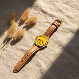 Puede incluir: Un reloj de pulsera con caja de metal dorado y correa de cuero color canela liso. La esfera es de color amarillo brillante y presenta una ilustraci&oacute;n gr&aacute;fica negra de tres pl&aacute;tanos de dibujos animados con gafas de sol y n&uacute;meros negros.