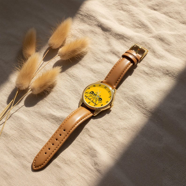 Puede incluir: Un reloj de pulsera con caja de metal dorado y correa de cuero color canela liso. La esfera es de color amarillo brillante y presenta una ilustraci&oacute;n gr&aacute;fica negra de tres pl&aacute;tanos de dibujos animados con gafas de sol y n&uacute;meros negros.