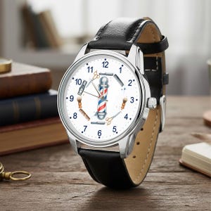 Peut inclure: Une montre-bracelet avec un bracelet en cuir noir et un boîtier argenté. Le cadran blanc présente un motif central de poteau de barbier entouré d'illustrations de blaireaux, de rasoirs droits et de ciseaux, avec des chiffres noirs.